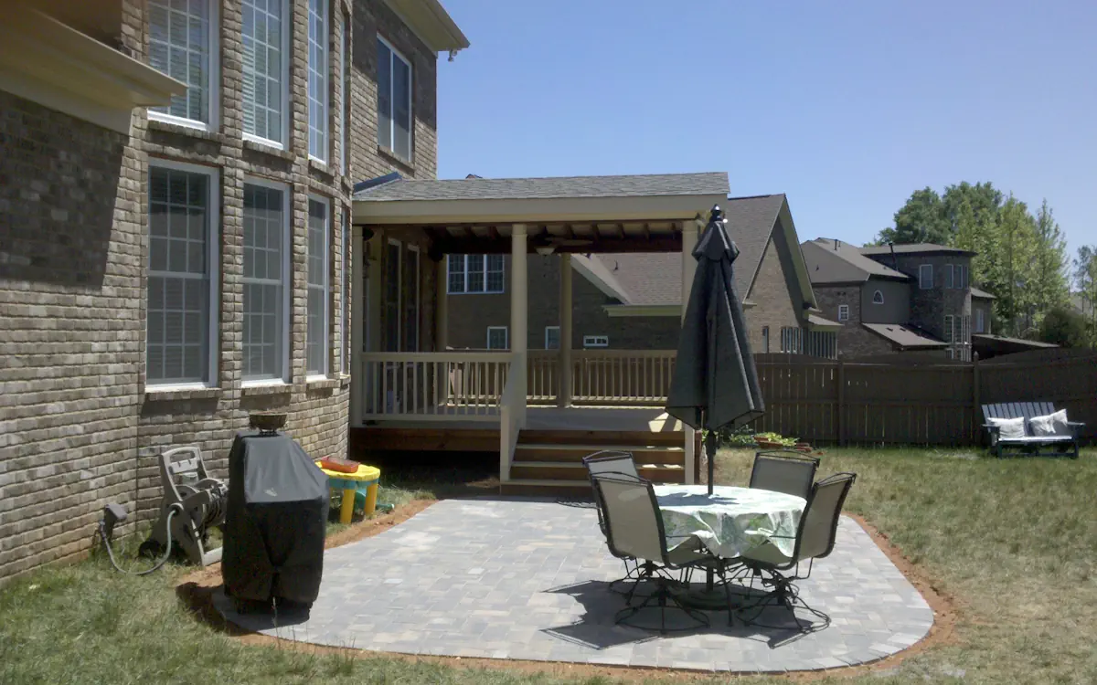 Light-colored columns, extending over a wooden deck with a railing. Steps lead down to a paved patio area, creating a connection between the porch and the backyard space.