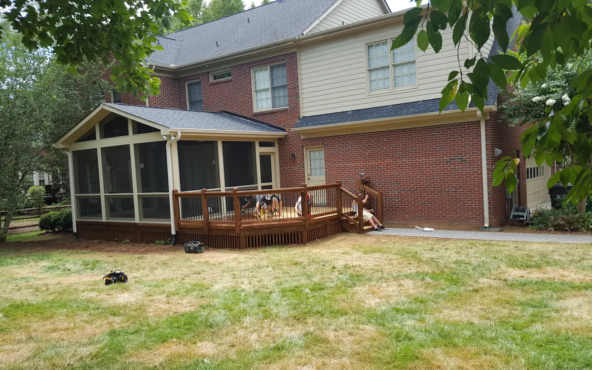 A two-story brick home features a beautifully crafted screened-in porch extending from the back, offering a shaded and protected outdoor space. The screen porch is enclosed with fine mesh screens and framed in soft beige, seamlessly blending with the house’s exterior. A gable roof with triangular screen openings enhances airflow, and a gutter system directs rainwater away from the structure. The screened porch is elevated and sits adjacent to a spacious wooden deck with a rich brown finish, complete with sturdy railings and built-in steps leading down to the yard. Beneath the screen porch, decorative slatted skirting adds a polished touch. In the background, lush greenery and neighboring homes provide a serene backdrop, while a group of people works together on the deck, possibly engaging in maintenance or improvement activities. The foreground showcases a lawn with mixed green and dry grass patches, adding a natural, lived-in feel to the backyard.