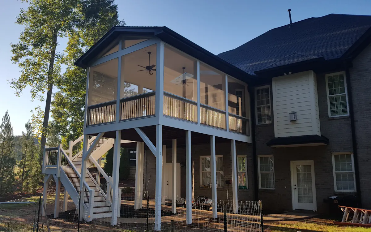 A modern two-story home features a striking elevated screened-in porch, designed to provide a breezy yet protected outdoor space. The screen porch is enclosed with fine mesh screens framed in white, ensuring an open feel while keeping insects out. A gable roof with exposed beams adds architectural character, while ceiling fans inside the screened porchenhance comfort. The structure is supported by tall white columns, creating a shaded area beneath the screened porchthat can be used for storage or additional outdoor activities. A wide staircase with white railings and black balusters leads up to the screen porch, seamlessly connecting it to the main house. The backyard features a partially fenced area, mature trees, and an expansive natural landscape in the background, adding to the home’s serene setting.