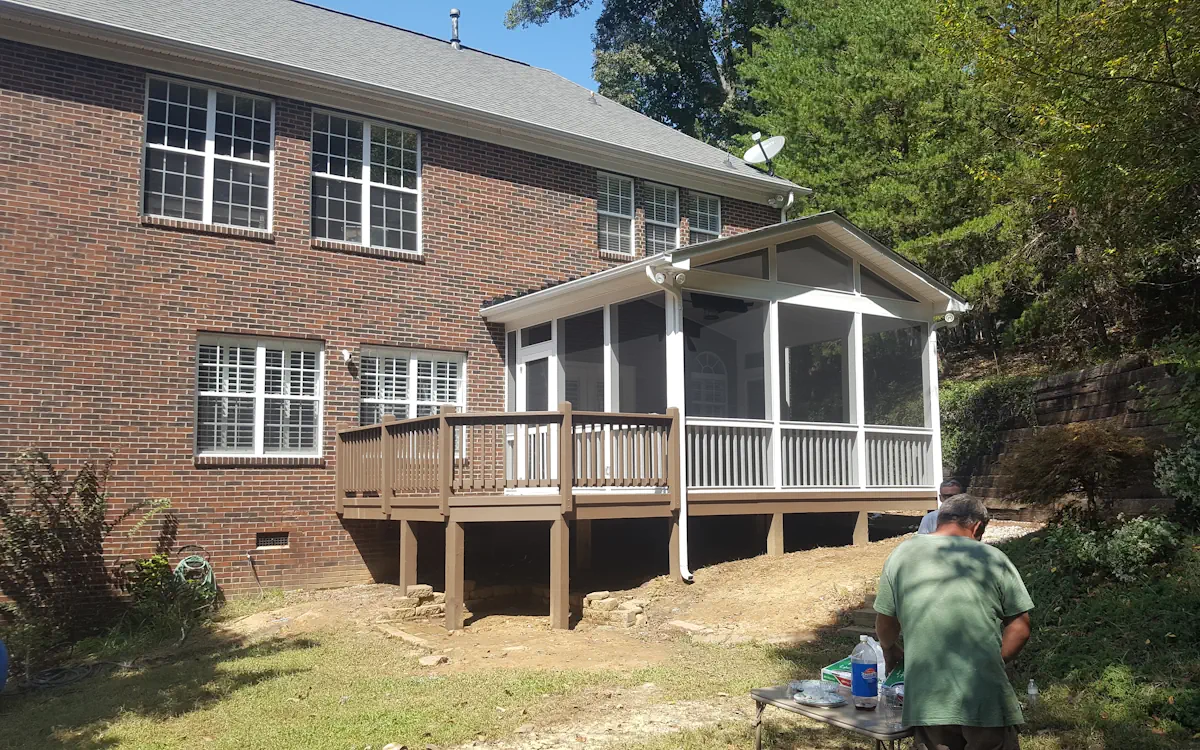 A two-story brick home features a screened-in porch extending from the back, offering a well-shaded and enclosed outdoor retreat. The screen porch is framed in white with fine mesh screens, allowing fresh air to circulate while keeping pests out. A gable roof with triangular screen openings enhances airflow, while a ceiling fan inside the screened porchprovides additional cooling. The structure is elevated and supported by sturdy posts, with a brown wooden deck extending from one side, creating an open-air transition between the screen porch and the backyard. Below the screened porch, the space is left open, providing ventilation and storage opportunities. The surrounding landscape includes a sloped yard, a retaining wall covered with greenery, and mature trees that add natural privacy. In the foreground, two people are engaged in an outdoor activity at a small folding table, indicating a relaxed backyard setting.