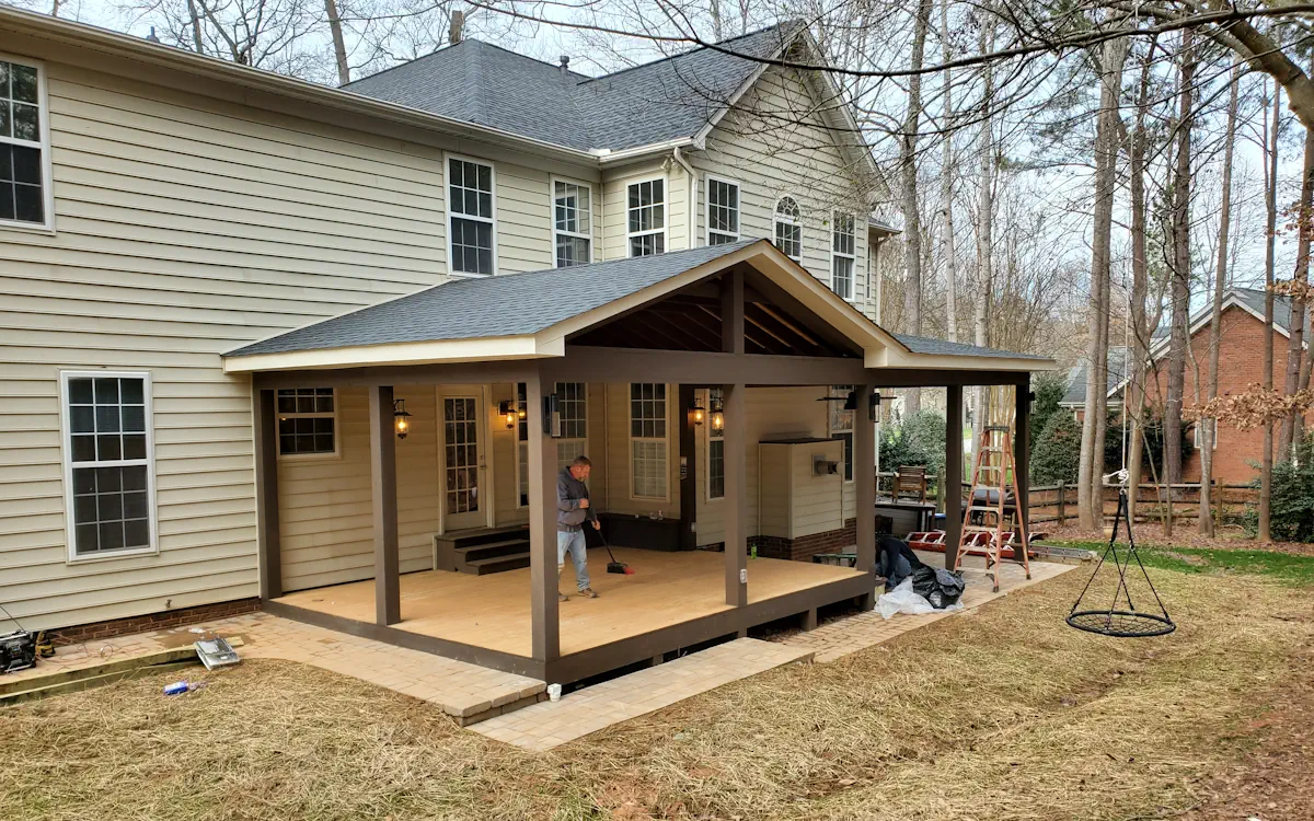 Covered porch with a gabled roof, sturdy square columns, and a spacious wooden deck. Warm wall-mounted lanterns complement the beige house siding, while a paved walkway connects the space to the yard.