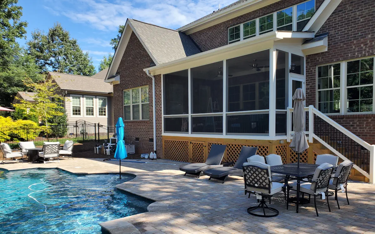 A luxurious brick home features a spacious screened-in porch overlooking a beautifully designed backyard with a pool. The screen porch is elevated and enclosed with fine mesh screens, framed in white, providing a shaded and protected outdoor retreat. Large windows above the screened porch allow natural light to filter in, while ceiling fans inside enhance airflow for added comfort. A set of stairs with white railings and black balusters leads from the screened porch to the patio area. Beneath the screened porch, a wooden lattice skirt encloses the space below for a polished appearance. The pool area is paved with elegant stone tiles and is furnished with cushioned lounge chairs, a dining table with an umbrella, and additional seating for relaxation. The surrounding landscape includes lush greenery, a neighboring house, and a black metal fence enclosing the yard.
