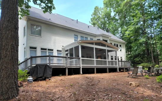 A large two-story home with white siding features an expansive screened-in porch at the rear, seamlessly integrating indoor and outdoor living. The screen porch is framed in white with fine mesh screens, allowing fresh air while keeping bugs out. A gable roof with exposed beams and triangular screen openings enhances ventilation, complementing the house’s modern yet classic design. The screened porch is elevated and connects to a spacious wooden deck with gray railings that wraps around the back of the home. Beneath the screen porch, an open area provides additional storage or shaded space. In the background, a wooded backyard with towering trees offers privacy, while a cozy seating area with outdoor chairs is arranged on the forest floor. A covered grill sits on the ground near the deck, suggesting a well-used outdoor entertainment space.