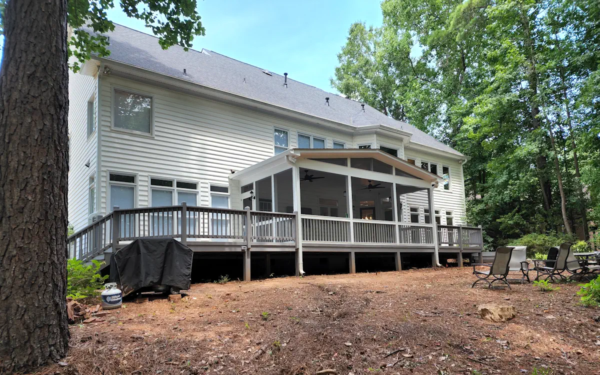A large two-story home with white siding features an expansive screened-in porch at the rear, seamlessly integrating indoor and outdoor living. The screen porch is framed in white with fine mesh screens, allowing fresh air while keeping bugs out. A gable roof with exposed beams and triangular screen openings enhances ventilation, complementing the house’s modern yet classic design. The screened porch is elevated and connects to a spacious wooden deck with gray railings that wraps around the back of the home. Beneath the screen porch, an open area provides additional storage or shaded space. In the background, a wooded backyard with towering trees offers privacy, while a cozy seating area with outdoor chairs is arranged on the forest floor. A covered grill sits on the ground near the deck, suggesting a well-used outdoor entertainment space.