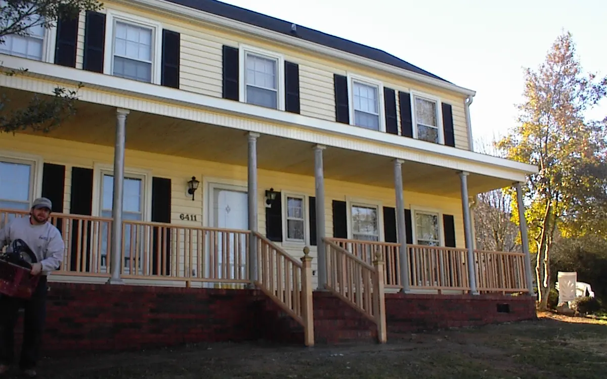 Porch cover extends across the front of the house, supported by evenly spaced round columns. A wooden railing with matching staircases leads to the entrance, while the brick foundation contrasts with the yellow siding and black shutters.