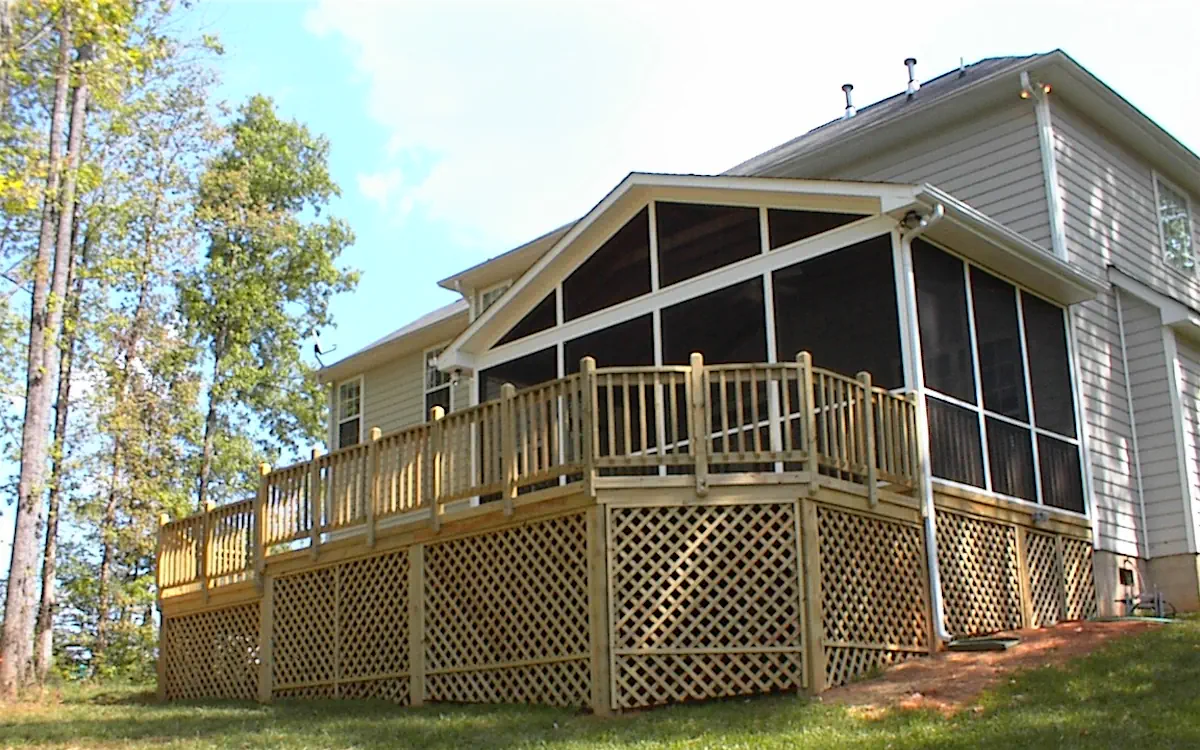 A two-story house with light gray siding features a screened-in porch attached to the back, providing a spacious and protected outdoor living area. The screen porch has a gable roof with white framing and large triangular screen openings at the top, allowing for better ventilation. The screened porch is fully enclosed with fine mesh screens, ensuring an insect-free outdoor experience. It is elevated above the ground and supported by wooden posts, with a decorative lattice skirt enclosing the underside. A wooden deck extends from the screen porch, featuring railings and a small staircase leading down to the yard. The surrounding area includes tall trees, a well-maintained lawn, and a natural wooded backdrop.