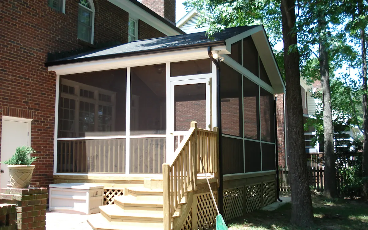 A two-story brick home features a screened-in porch attached to the back, offering a protected outdoor space. The screen porch has a gable roof with white trim and fine mesh screens, allowing for fresh air while keeping insects out. A wooden staircase with railings leads up to the screened porch, which has a screened door for entry. The lower section of the screen porch is enclosed with wooden lattice skirting. The space around the screened porch includes a paved patio area with a storage box and a potted plant. Mature trees surround the property, providing shade, and a wooden picket fence is visible in the background.