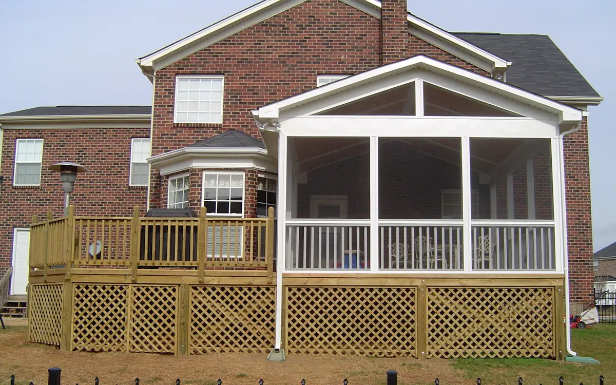 A two-story brick home features a screened-in porch attached to the back, providing a comfortable and enclosed outdoor space. The screen porch has a gable roof with white framing and fine mesh screens, ensuring protection from insects while maintaining airflow. The lower portion of the screened porch includes a railing for safety, and its structure is elevated, supported by a wooden frame. Below the screen porch, a decorative lattice skirt encloses the underside, complementing the adjacent wooden deck. The deck extends to the left, featuring a railing, built-in grill area, and a patio heater. The surrounding yard is mostly bare with patches of grass, and neighboring houses are visible in the background under an overcast sky.