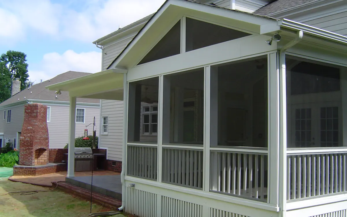 A light-colored two-story home features a screened-in porch attached to the back, providing a protected and shaded outdoor living area. The screen porch has a gable roof with white framing and fine mesh screens, ensuring an airy yet insect-free environment. Large triangular screen openings at the top of the screened porch allow for additional ventilation. The lower portion of the screen porch has a railing for safety, and a decorative skirting encloses the space beneath. To the left of the screen porch, a covered patio extends outward, featuring a built-in outdoor kitchen with a stainless steel grill and a red brick fireplace. The backyard is partially landscaped, with neighboring houses visible in the background under a partly cloudy sky.