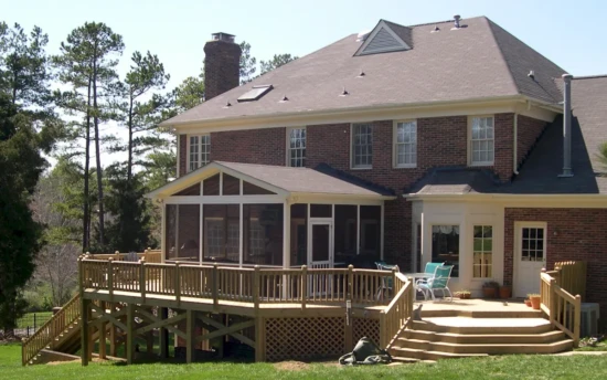 A large two-story brick home features an elevated screened-in porch extending from the back, providing a comfortable and enclosed outdoor space. The screen porch is framed in white with fine mesh screens, allowing fresh air while keeping bugs out. A gable roof with triangular screen openings enhances ventilation, and a screened door provides access to the spacious screen porch. Beneath the screened porch, a wooden lattice skirt encloses the support structure, blending seamlessly with the adjacent elevated wooden deck. The deck extends outward with built-in railings and multiple levels, creating an expansive outdoor entertainment area. A wide staircase leads down to a well-manicured lawn, and to the right of the screen porch, a separate patio area is furnished with outdoor chairs and potted plants. The property is surrounded by tall trees, providing shade and a natural backdrop.
