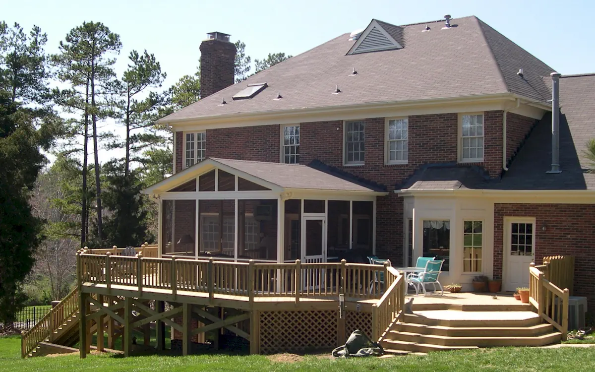 A large two-story brick home features an elevated screened-in porch extending from the back, providing a comfortable and enclosed outdoor space. The screen porch is framed in white with fine mesh screens, allowing fresh air while keeping bugs out. A gable roof with triangular screen openings enhances ventilation, and a screened door provides access to the spacious screen porch. Beneath the screened porch, a wooden lattice skirt encloses the support structure, blending seamlessly with the adjacent elevated wooden deck. The deck extends outward with built-in railings and multiple levels, creating an expansive outdoor entertainment area. A wide staircase leads down to a well-manicured lawn, and to the right of the screen porch, a separate patio area is furnished with outdoor chairs and potted plants. The property is surrounded by tall trees, providing shade and a natural backdrop.