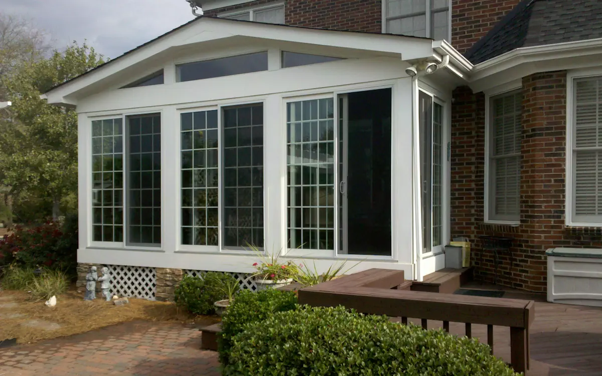 Gabled sunroom with a white aluminum frame, large grid-patterned sunroom windows, transom windows for extra light, and a sliding glass door for easy access.