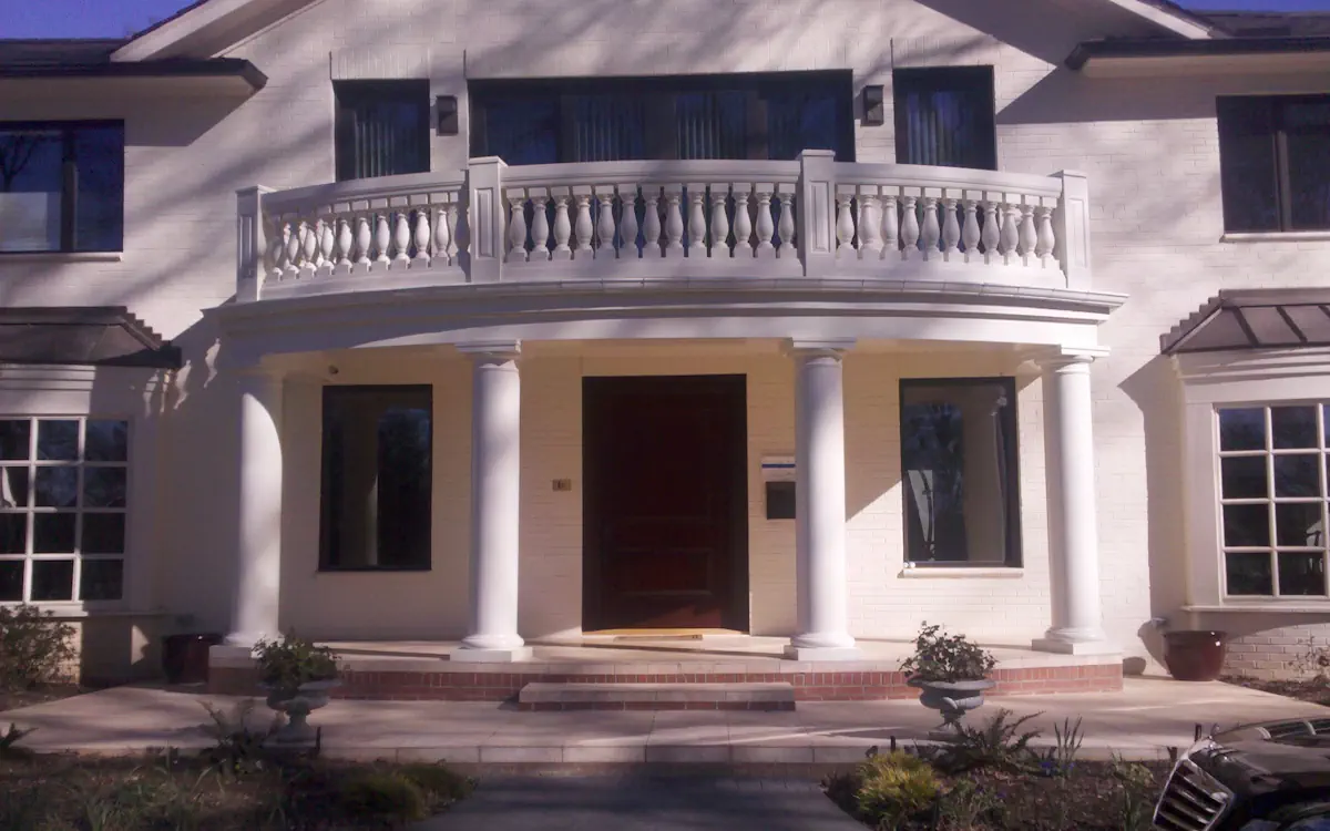 Porch cover features a curved roof supported by white round columns, matching the home's exterior. A decorative balustrade above adds a classic architectural element. Brick steps lead to the entrance, framed by large windows on either side.