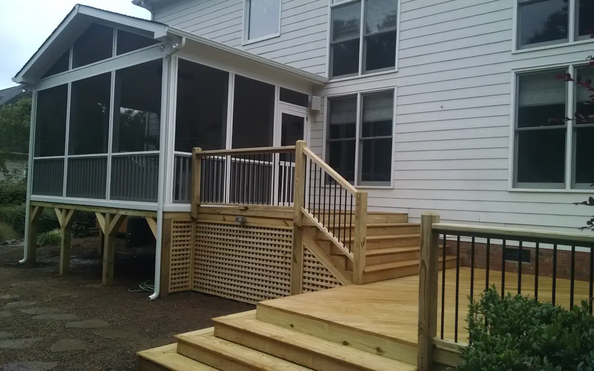 A two-story house with white siding features a screened-in porch attached to the back, providing a protected outdoor living space. The screen porch is elevated, supported by wooden posts, and enclosed with fine mesh screens framed in white, allowing for ventilation while keeping insects out. The screened porch has a gable roof with triangular screen openings at the top. A set of wooden stairs with black metal balusters connects the screen porch to a large wooden deck, which extends into the backyard. The deck features multiple levels with railings and a lattice skirt enclosing the area beneath the screen porch. The surrounding landscape includes gravel pathways, shrubs, and a well-maintained lawn.