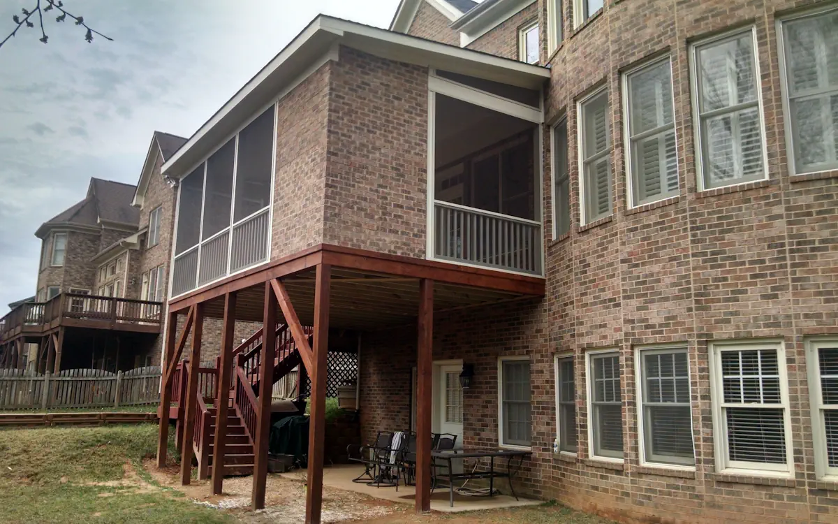 A large brick home features a screened-in porch elevated on sturdy wooden posts, creating a spacious covered area underneath. The screen porch is enclosed with fine mesh screens and white framing, providing a comfortable, protected outdoor living space. A set of wooden stairs with railings leads up to the screened porch, which includes a railing along the lower portion for added safety. Beneath the screen porch, a patio area is furnished with a dining table and chairs, offering an additional outdoor gathering space. Neighboring homes with similar architectural styles and wooden decks are visible in the background. The surrounding yard has patches of grass and a wooden fence lining the property.