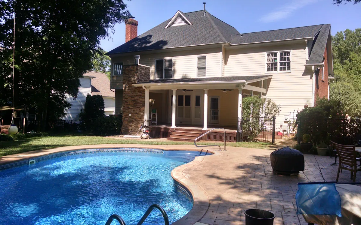 A flat roof extends from the house over a wooden deck, supported by white round columns. A stone chimney serves as a structural feature, while ceiling fans enhance airflow. This covered porch overlooks a pool and a paved patio area.