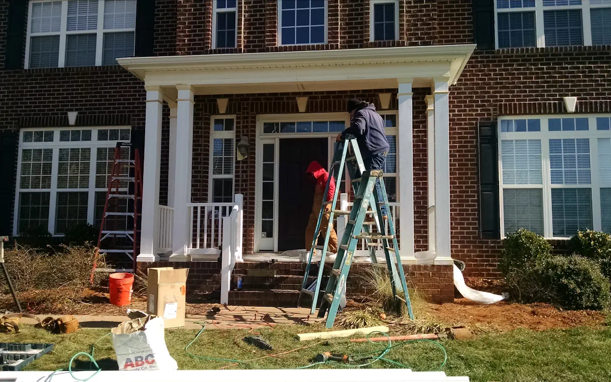 Covered porch features a flat roof supported by white square columns, complementing the brick exterior. A white railing encloses one side, and the porch extends over the front entrance, providing shade and protection.