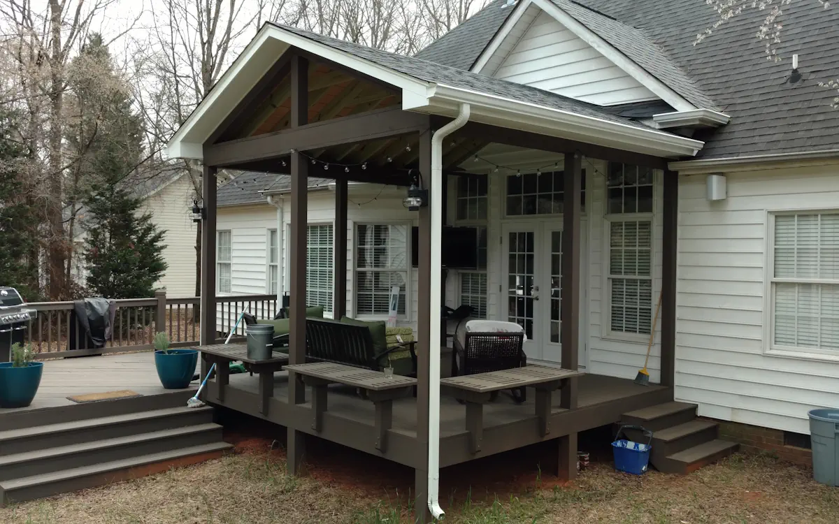 Exposed beams rest on dark brown columns, extending over an elevated wooden deck. Built-in benches, outdoor seating, and a perimeter railing define the space. Porch cover also includes string lights and a mounted TV for added functionality.