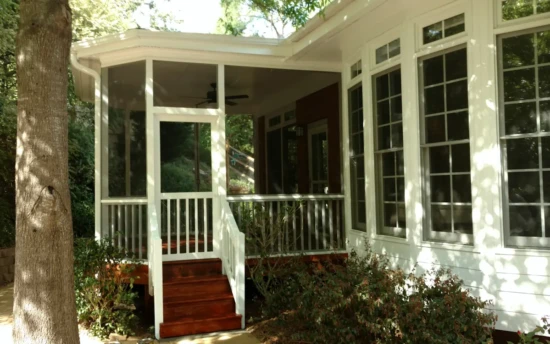 A house with a screened porch featuring white framing and fine mesh screens, providing a comfortable and protected outdoor space. The screen porch is elevated, with a set of rich brown wooden steps leading up to a white railing and a screened door. A ceiling fan is visible inside the porch, enhancing air circulation. Large windows with white trim line the exterior of the house, allowing natural light to enter. The surrounding area includes lush greenery, a tree trunk in the foreground, and bushes growing near the base of the porch. Sunlight filters through the trees, casting dappled shadows on the house and porch.