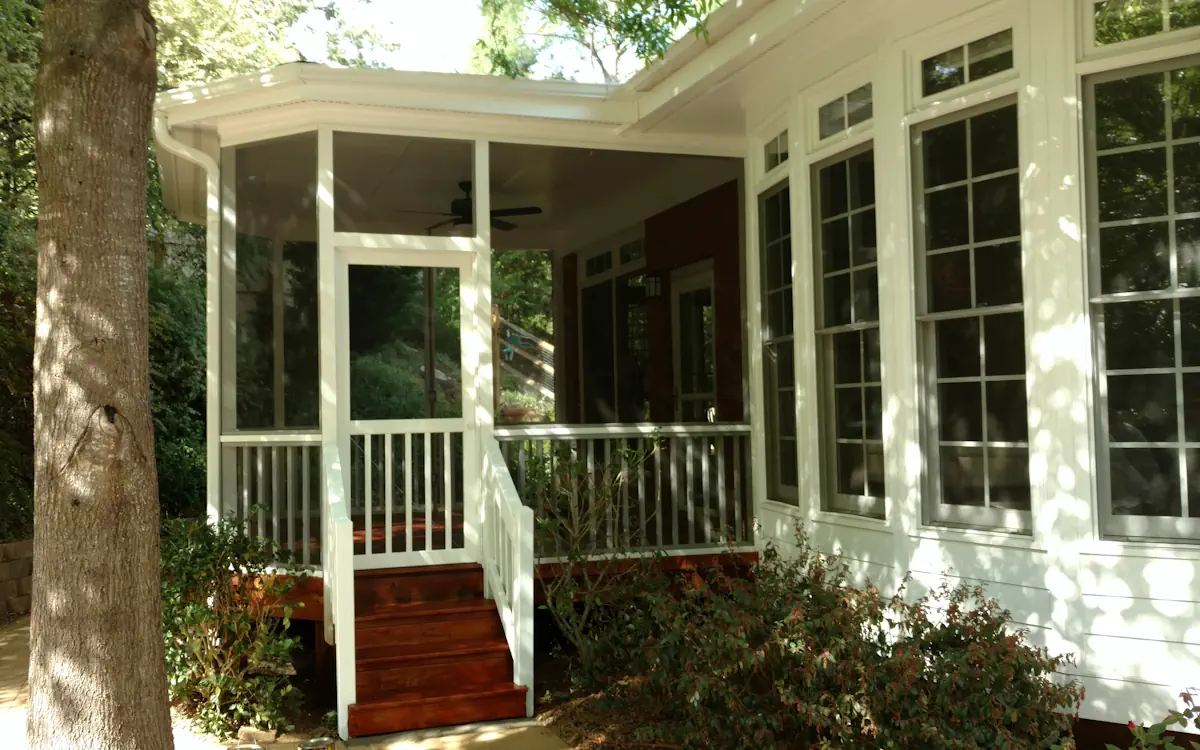 A house with a screened porch featuring white framing and fine mesh screens, providing a comfortable and protected outdoor space. The screen porch is elevated, with a set of rich brown wooden steps leading up to a white railing and a screened door. A ceiling fan is visible inside the porch, enhancing air circulation. Large windows with white trim line the exterior of the house, allowing natural light to enter. The surrounding area includes lush greenery, a tree trunk in the foreground, and bushes growing near the base of the porch. Sunlight filters through the trees, casting dappled shadows on the house and porch.