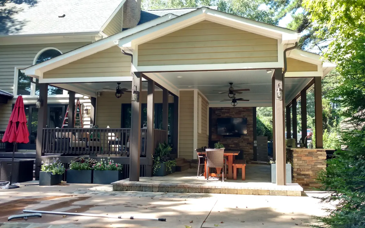 Porch cover features a gabled roof supported by dark brown columns, matching the beige siding. The space includes a seating area with a railing, planter boxes, and an outdoor dining section with a stone fireplace and mounted TV.