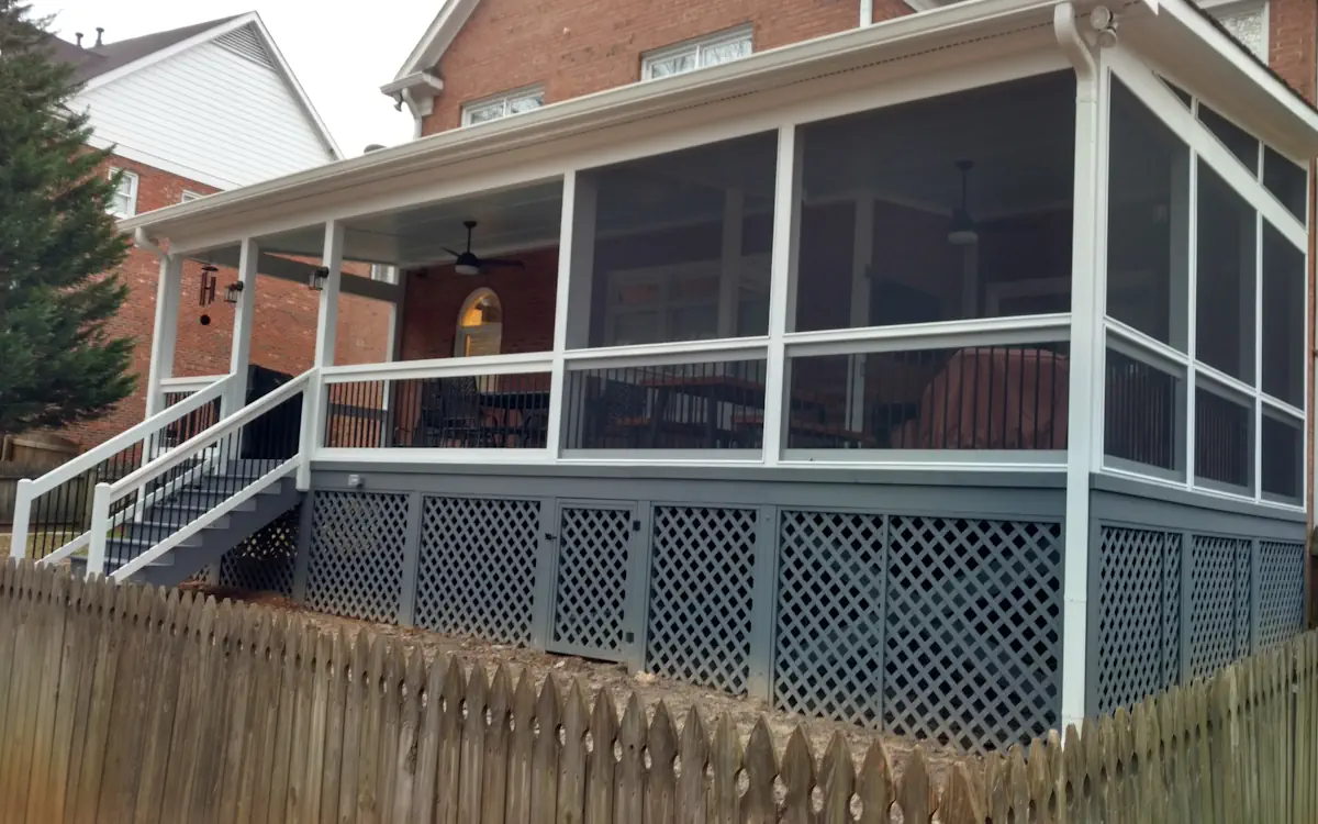 A two-story brick house with a screened-in porch attached to the back, elevated above the ground. The porch features white framing and fine mesh screens, enclosing a spacious outdoor living area furnished with a dining table and chairs. A ceiling fan is visible inside for added comfort. The structure is supported by a gray lattice skirt, which provides a decorative and functional enclosure beneath the screen porch. A set of stairs with white railings and black balusters leads up to the screened area. A wooden picket fence runs along the bottom of the image, enclosing the yard. The background includes neighboring houses and trees.