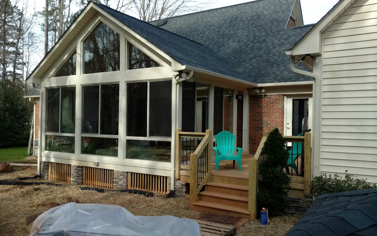 White sunroom with a gabled roof, large sunroom windows with triangular transoms, solid lower panels with stone accents, and an integrated entryway to the adjacent deck.