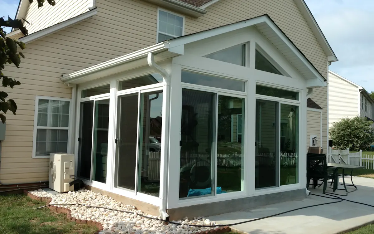 White sunroom with large sunroom windows, triangular transoms, sliding glass doors for entry, and a concrete foundation with decorative stone edging.