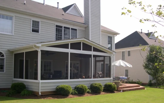 A two-story suburban home with light gray siding features a screen porch attached to the back. The screened-in porch has a gable roof with white framing and fine mesh screens, allowing for an open yet protected outdoor space. Inside the porch, a ceiling fan and various seating arrangements are visible, providing a comfortable setting for relaxation. The structure is slightly elevated, with neatly trimmed bushes lining the base. To the right of the screen porch, a small brick patio with a round table and a white patio umbrella extends into the yard. Neighboring houses and greenery surround the property, enhancing the inviting outdoor space.