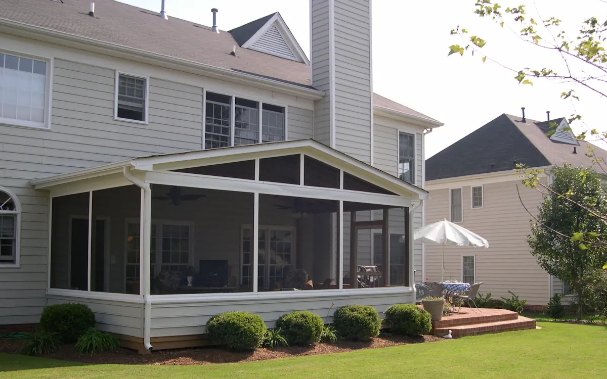 A two-story suburban home with light gray siding features a screen porch attached to the back. The screened-in porch has a gable roof with white framing and fine mesh screens, allowing for an open yet protected outdoor space. Inside the porch, a ceiling fan and various seating arrangements are visible, providing a comfortable setting for relaxation. The structure is slightly elevated, with neatly trimmed bushes lining the base. To the right of the screen porch, a small brick patio with a round table and a white patio umbrella extends into the yard. Neighboring houses and greenery surround the property, enhancing the inviting outdoor space.