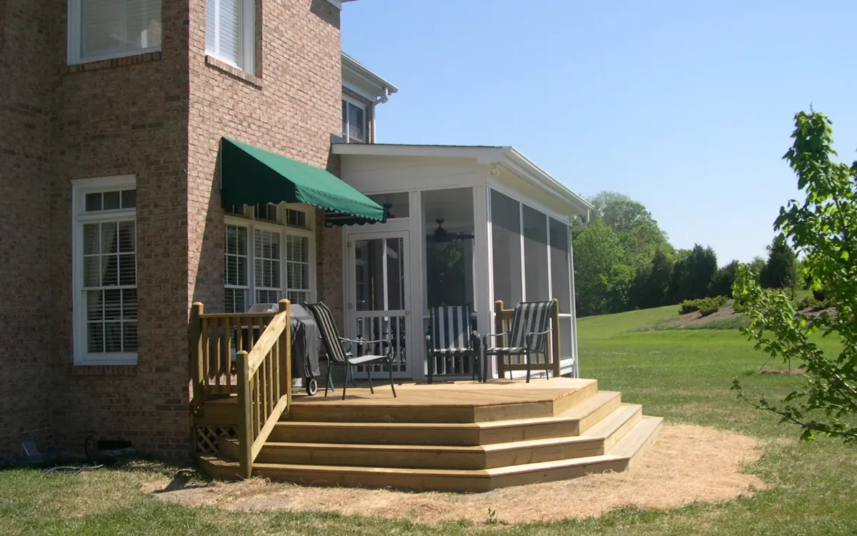 A brick two-story house with a screened-in porch attached to the back, featuring white framing and mesh screens that provide a protected outdoor space. The porch has a sloped roof and a ceiling fan inside. Adjacent to the porch is a newly built wooden deck with multiple wide steps leading down to the grassy yard. The deck is furnished with striped patio chairs and a grill. A green awning shades a small section of the house's exterior windows. The background consists of an open lawn with scattered trees and shrubs under a clear blue sky.