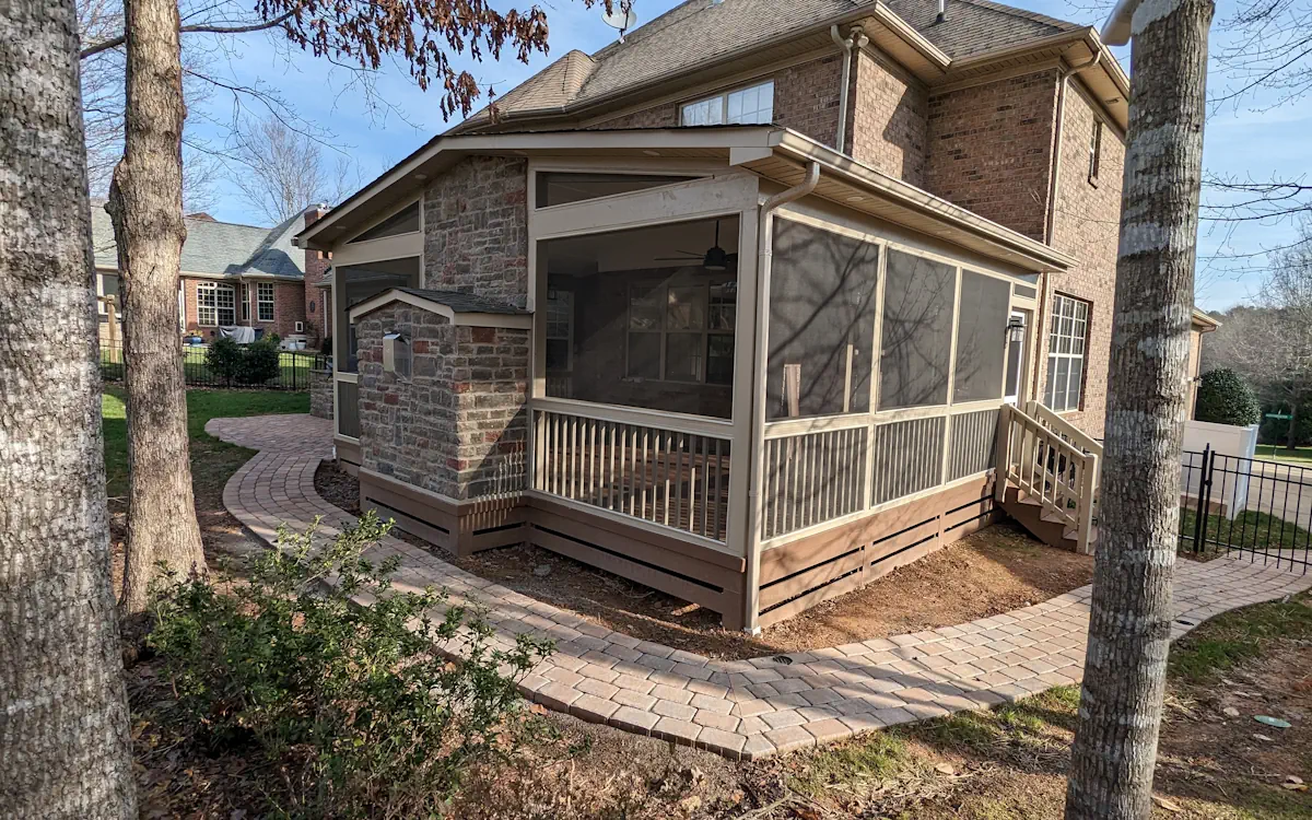 A two-story brick house with a screened-in porch attached to the back, featuring beige framing and fine mesh screens that provide a protected outdoor space. The porch is elevated, with wooden railings and a set of stairs leading down to a paved walkway made of interlocking bricks that curves around the yard. A ceiling fan is visible inside the porch, along with large windows offering a view of the house’s interior. The exterior combines brick and stone accents, adding a rustic charm. The surrounding yard features trees, shrubs, and a neighboring house in the background.