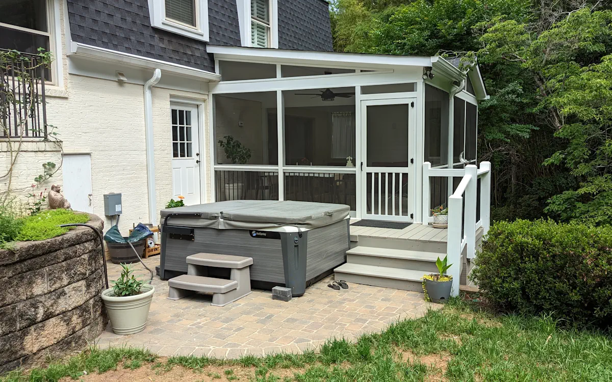 A two-story house with white brick and dark gray siding features a screened-in porch attached to the back. The porch has a modern design with a sloped roof, white framing, and fine mesh screens, creating a protected outdoor space. A ceiling fan is visible inside, along with patio furniture. The porch is accessed by a small set of gray stairs with white railings, leading down to a paved patio area. In the foreground, a large gray hot tub with a matching cover and small steps is situated on the patio. The surrounding area includes greenery, potted plants, a retaining wall with landscaping, and a dense tree line in the background.