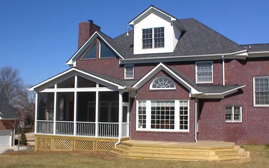 A two-story brick home features a screened-in porch attached to the back, providing a spacious and protected outdoor living area. The screen porch is elevated, with white framing and fine mesh screens enclosing the space while allowing for airflow. A gable roof with triangular screen openings enhances the screened porch design, complementing the home's architectural style. Below the screen porch, a wooden lattice skirt encloses the underside. A white downspout runs along the side, directing rainwater away from the foundation. Adjacent to the screen porch, a large wooden deck with multiple steps leads to the backyard, offering an additional outdoor gathering area. The background includes neighboring homes, trees, and a clear blue sky.