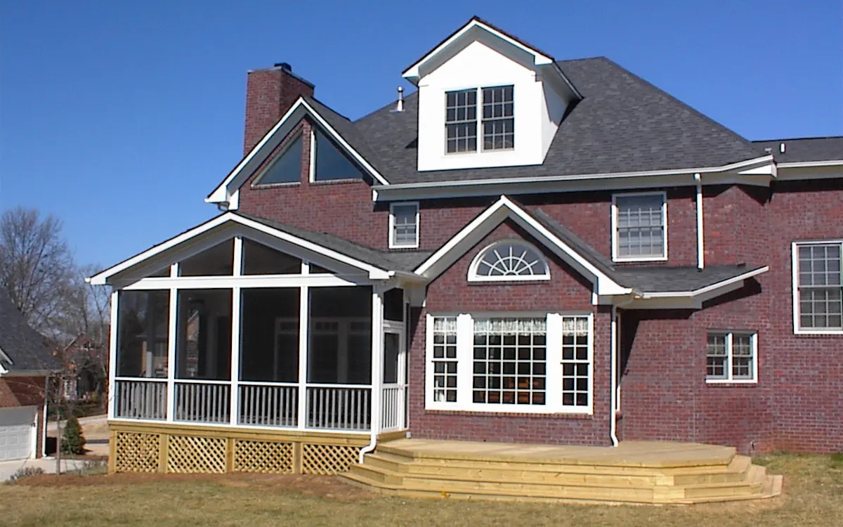 A two-story brick home features a screened-in porch attached to the back, providing a spacious and protected outdoor living area. The screen porch is elevated, with white framing and fine mesh screens enclosing the space while allowing for airflow. A gable roof with triangular screen openings enhances the screened porch design, complementing the home's architectural style. Below the screen porch, a wooden lattice skirt encloses the underside. A white downspout runs along the side, directing rainwater away from the foundation. Adjacent to the screen porch, a large wooden deck with multiple steps leads to the backyard, offering an additional outdoor gathering area. The background includes neighboring homes, trees, and a clear blue sky.