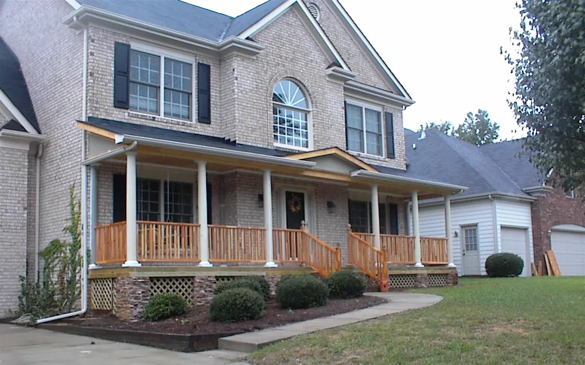 A wraparound design extends across the front of the house, supported by white round columns. The wooden railing and steps contrast with the brick exterior, while lattice skirting adds detail below. Covered porch provides a sheltered entryway and additional outdoor space.