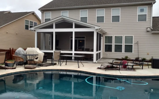 A modern two-story home with light beige siding features a screened-in porch extending from the back, offering a comfortable and shaded outdoor space. The screen porch is enclosed with fine mesh screens and framed in white, allowing for fresh air circulation while providing protection from insects. A gable roof with triangular screen openings enhances airflow, and a ceiling fan inside the screened porch ensures additional comfort. The screen porch seamlessly connects to a backyard pool area, which is surrounded by a paved patio with multiple lounge chairs, a dining set, and covered grills. The pool’s blue water reflects the surrounding outdoor elements, adding to the inviting atmosphere. Neighboring houses with similar architectural styles can be seen in the background, along with a wooden fence providing a sense of privacy. The backyard is arranged for relaxation and outdoor gatherings, making the screened porch a natural extension of the home's entertainment space.