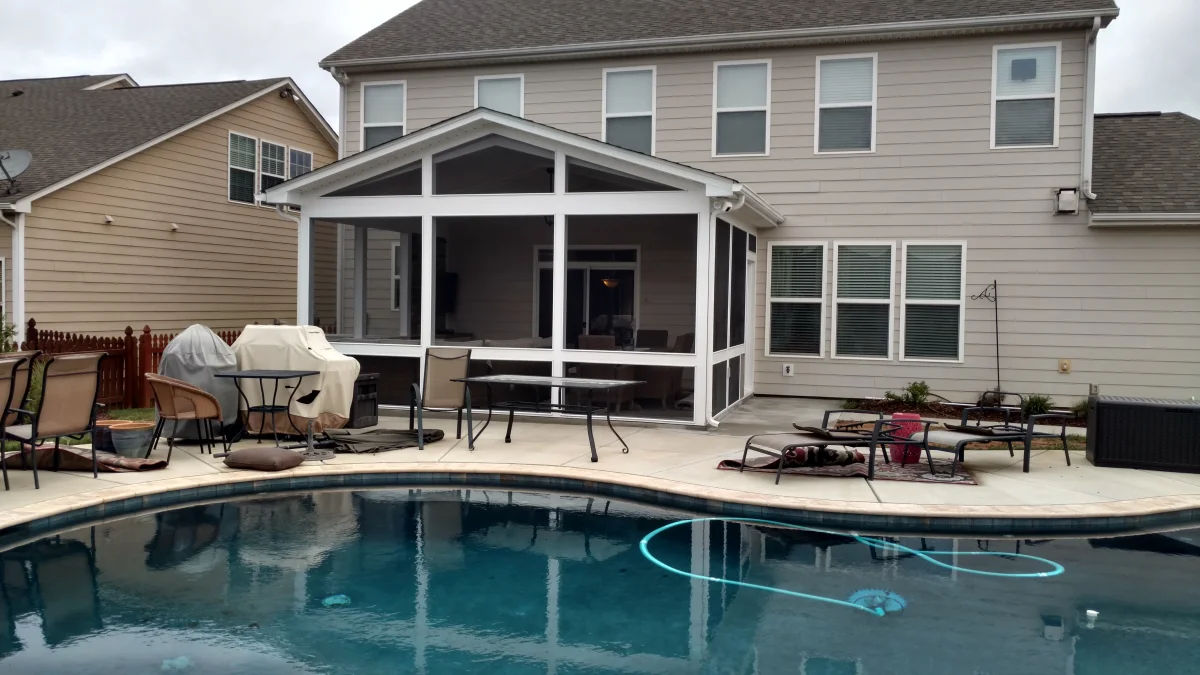 A modern two-story home with light beige siding features a screened-in porch extending from the back, offering a comfortable and shaded outdoor space. The screen porch is enclosed with fine mesh screens and framed in white, allowing for fresh air circulation while providing protection from insects. A gable roof with triangular screen openings enhances airflow, and a ceiling fan inside the screened porch ensures additional comfort. The screen porch seamlessly connects to a backyard pool area, which is surrounded by a paved patio with multiple lounge chairs, a dining set, and covered grills. The pool’s blue water reflects the surrounding outdoor elements, adding to the inviting atmosphere. Neighboring houses with similar architectural styles can be seen in the background, along with a wooden fence providing a sense of privacy. The backyard is arranged for relaxation and outdoor gatherings, making the screened porch a natural extension of the home's entertainment space.