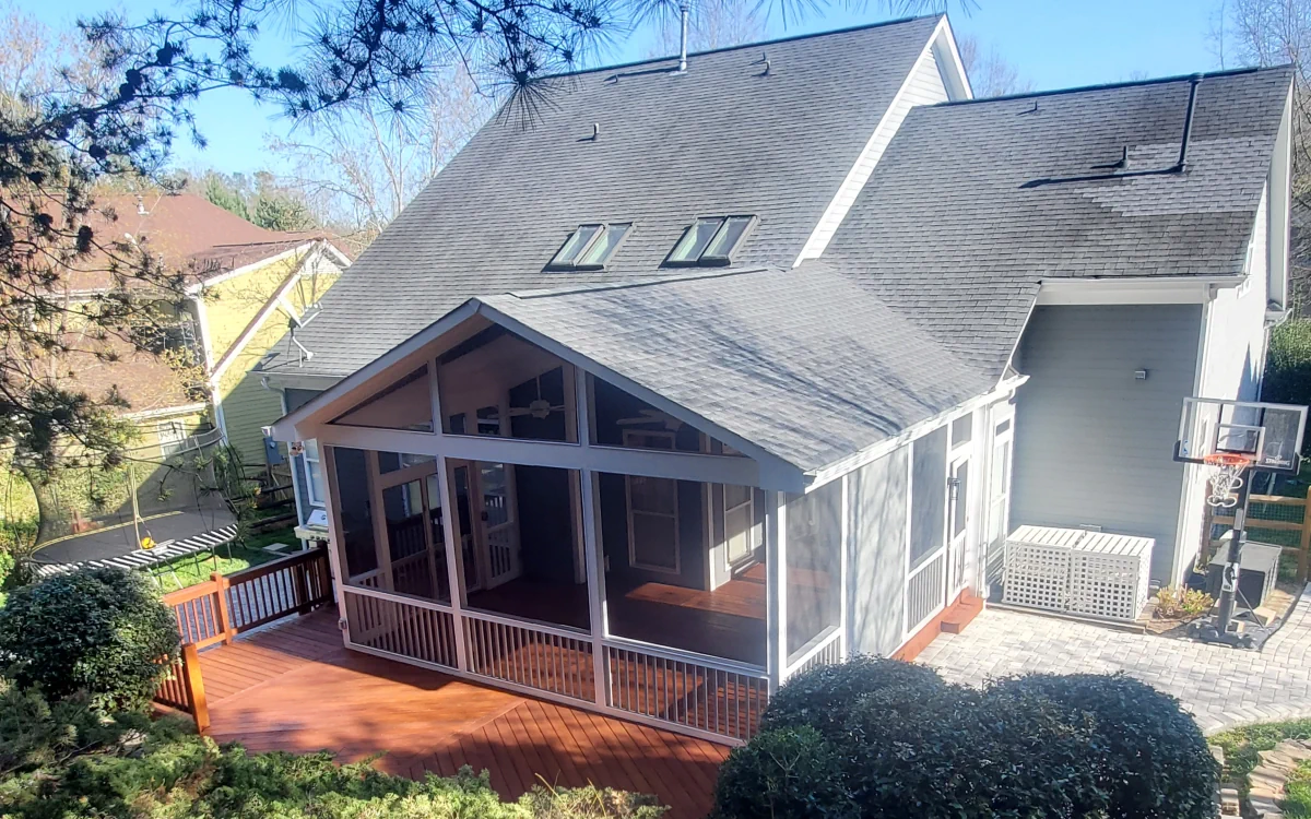 A two-story suburban house with a steeply pitched dark gray shingled roof, featuring multiple skylights. Attached to the back of the house is a screened-in porch, enclosed with fine mesh screens framed by white trim, allowing for an open yet protected outdoor space. The screen porch has a gable roof and a wooden floor that extends into a spacious reddish-brown deck. Adjacent to the porch is a small paved patio with a white lattice-enclosed outdoor unit. A basketball hoop is mounted on a pole near the patio. In the background, neighboring houses, trees, and a trampoline with a safety net are visible. Sunlight casts shadows, highlighting the structure and design of the screened porch.