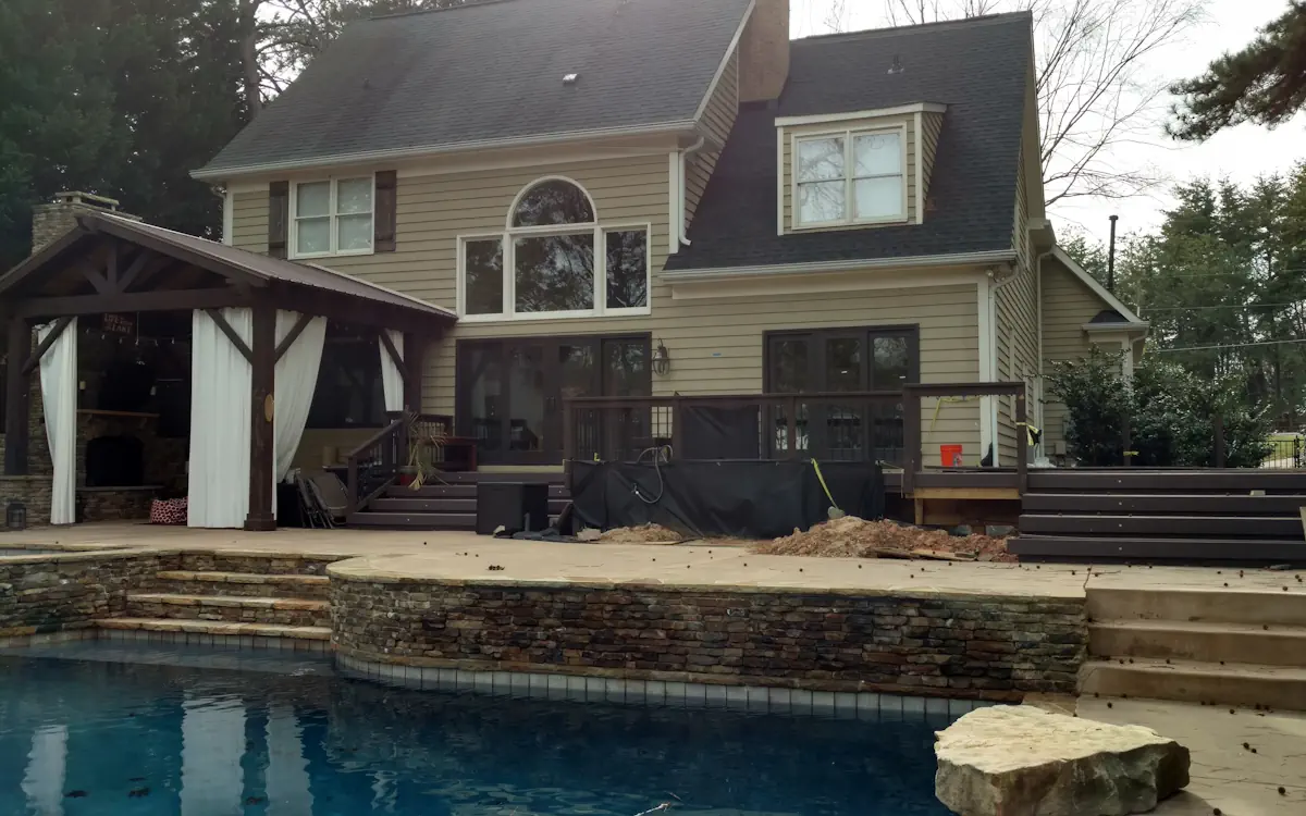 Outdoor kitchen set under a rustic wooden pavilion with exposed beams and white curtains for a cozy, private feel. A built-in stone fireplace adds warmth and ambiance. The space is connected to a multi-level deck overlooking a swimming pool, with stone steps and retaining walls enhancing the natural look.
