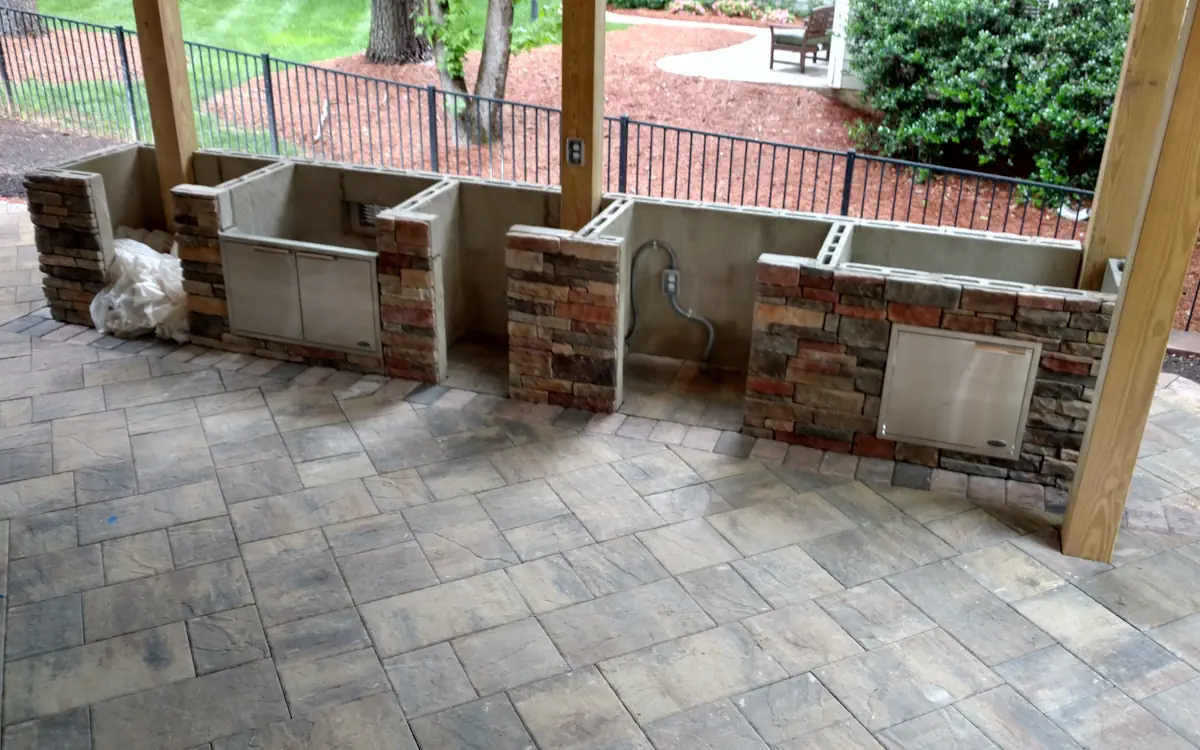 Partially built outdoor kitchen with a stone and stucco base, stainless steel storage doors, and electrical and gas line preparations. Covered space with stylish paver flooring, built for grilling and entertaining.