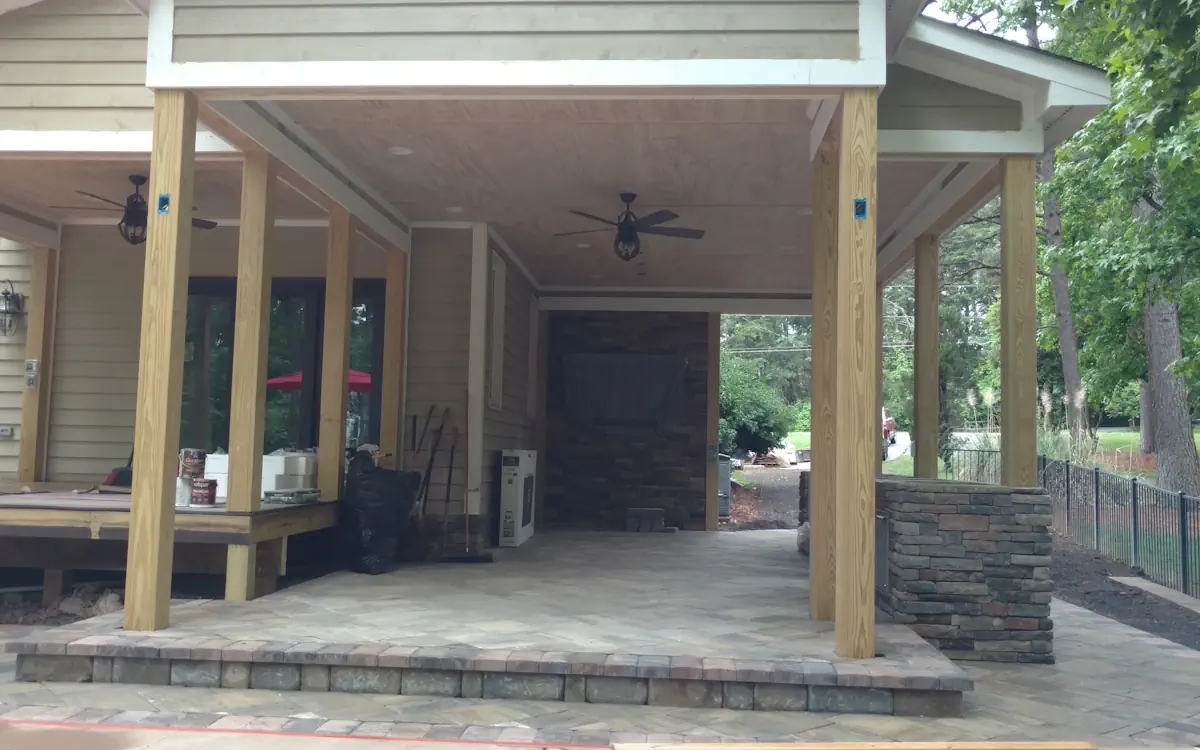 Spacious covered outdoor kitchen under construction, featuring sturdy wooden posts, a stone-accented wall, and a high ceiling with fans.