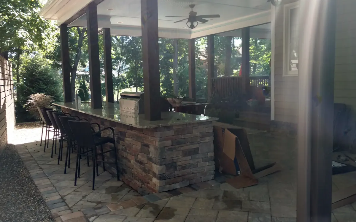Stylish outdoor kitchen with a stone and granite bar, built-in grill, and sink. High-top seating, wooden posts, and a covered ceiling with fans create a comfortable and inviting space for outdoor dining and entertaining.