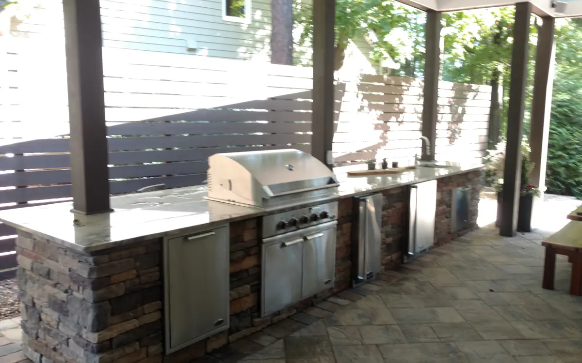 Outdoor kitchen with a built-in stainless steel grill, under-counter refrigerators, and a sink. Countertop features stone and granite for a stylish and durable workspace. Covered design and modern privacy fencing.