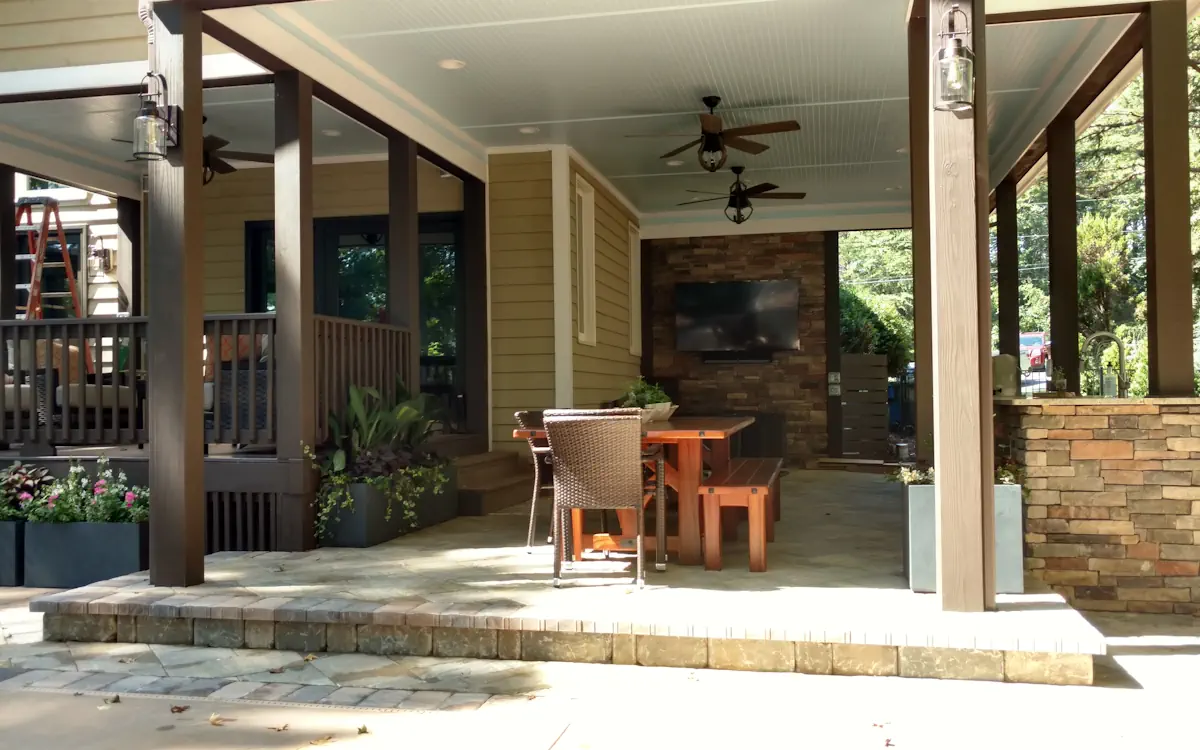 Elegant outdoor kitchen and dining space with a stone accent wall, mounted TV, and wooden picnic-style table. Covered ceiling with fans, stylish lighting, and lush greenery.